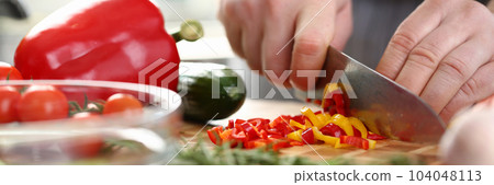 Hands of cook cut yellow and red peppers on cutting board with other vegetables on table in kitchen 104048113