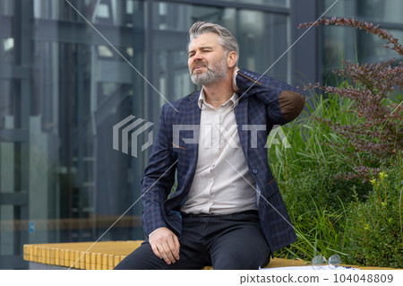 Overtired mature man resting on bench outside office building after work, businessman having severe neck pain, gray-haired boss in suit massaging muscle on lunch break. 104048809