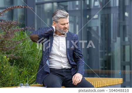 Overtired mature man resting on bench outside office building after work, businessman having severe neck pain, gray-haired boss in suit massaging muscle on lunch break. Overtired mature man resting on bench outside office building after work, businessman having severe neck pain, gray-haired boss in suit massaging muscle on lunch break. 104048810