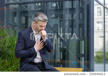 Sick mature man outside office building, adult gray-haired businessman coughing sitting on bench near work, senior boss in business suit with cold virus. 104048812