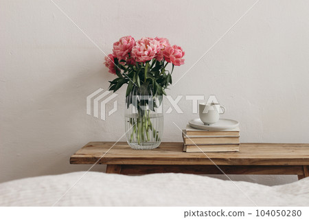 Feminine interior, still life. Rippled glass vase with coral pink peony flowers. Floral arrangement. Cup of coffee, books on vintage teak wooden bench, table. Empty white wall background. Blurred bed 104050280