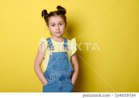 Beautiful smart clever charming little child girl, adorable school kid, pupil in yellow t-shirt and jeans overalls, holding hands in pockets, confidently looking at camera, isolated yellow background 104059784