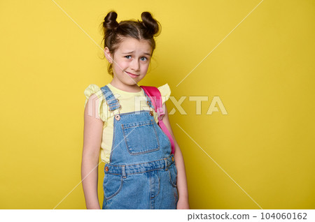 Caucasian emotional doubtful little kid girl 6 years old, primary school student, pupil with pink backpack, dressed in casual blue denim and yellow t-shirt, isolated on yellow color studio background 104060162