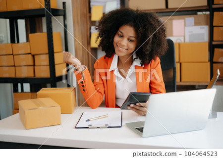 Startup small business SME, Entrepreneur owner African woman using smartphone or tablet taking receive and checking online purchase shopping order to preparing pack product box. .. 104064523