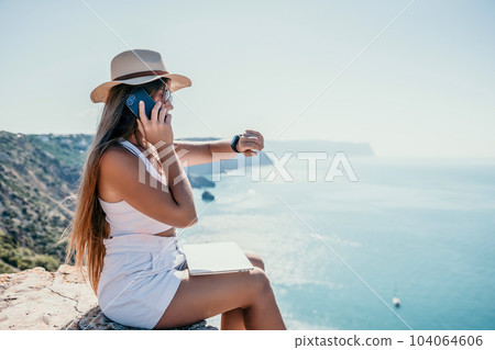 Digital nomad, woman in the hat, a business woman with a laptop sits on the rocks by the sea during sunset, makes a business transaction online from a distance. Freelance, remote work on vacation. Digital nomad, woman in the hat, a business woman with a laptop sits on the rocks by the sea during sunset, makes a business transaction online from a distance. Freelance, remote work on vacation. 104064606