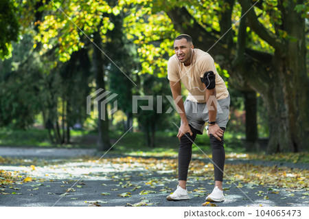 Tired man resting in park after jogging and fitness class, hispanic man using phone listening to music and podcasts in headphones breathing tired and exhausted. 104065473