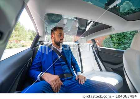 A caucasian man in a blue suit sits in the back seat of a car. Business class passenger.  104065484