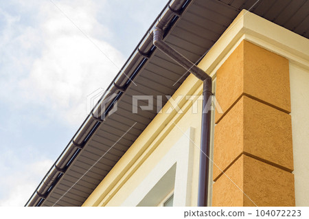 Close-up detail of cottage house corner with brown metal planks siding and roof with steel gutter rain system. Roofing, construction, drainage pipes installation and connection concept. Close-up detail of cottage house corner with brown metal planks siding and roof with steel gutter rain system. Roofing, construction, drainage pipes installation and connection concept. 104072223