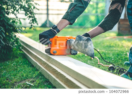 The man grinds the boards with a belt grinder, the electric tool for grinding and polishing the surface of the wooden board. The man grinds the boards with a belt grinder, the electric tool for grinding and polishing the surface of the wooden board. 104072966