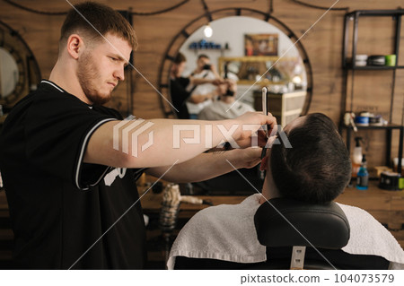 Barber using razor to shave beard for customer. Confident guy sitting at beauty salon Barber using razor to shave beard for customer. Confident guy sitting at beauty salon 104073579