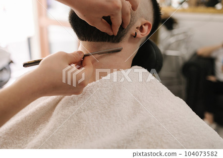 First person view of barber using razor to shave beard for customer. Confident guy sitting at beauty salon 104073582