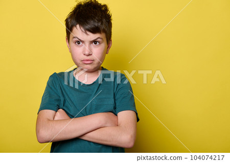 Isolated close-up emotional portrait on yellow background of a teenager boy, elementary school student with arms folded, looking at camera with sad, unhappy and sorrowful gaze. Back to school. 104074217