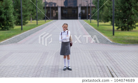 A sad caucasian girl in uniform and with a backpack holds her arms crossed on her chest stands outdoors after school. A sad caucasian girl in uniform and with a backpack holds her arms crossed on her chest stands outdoors after school. 104074689