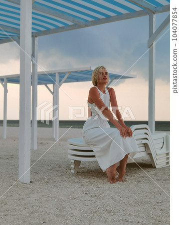 Portrait of blond young woman on empty beach wearing white dress and summer hat. Portrait of blond young woman on empty beach wearing white dress and summer hat. 104077854