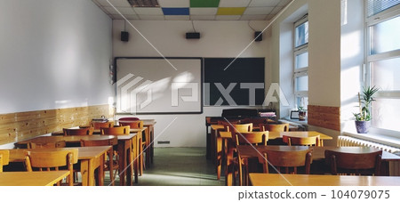 Music solfeggio empty class at school. The sun's rays fall on the floor through the window. Wooden student tables and chairs. School board and white walls. School interior Music solfeggio empty class at school. The sun's rays fall on the floor through the window. Wooden student tables and chairs. School board and white walls. School interior 104079075
