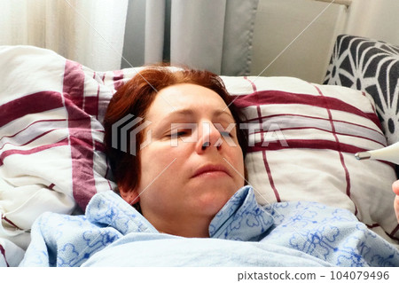 Sick woman in bed. Headache, runny nose and cold. Paleness of the face and faintness of the body. Feeling unwell. The patient lies on a pillow, holds a thermometer and measures the temperature 104079496