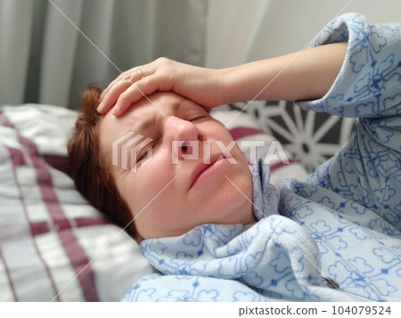 Sick woman in bed. Headache, runny nose and cold. Paleness of the face and faintness of the body. Feeling unwell. The patient lies on a pillow, holding forehead with his hand. Sharp pain and grimace. 104079524