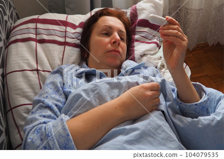 Sick woman in bed. Headache, runny nose and cold. Paleness of the face and faintness of the body. Feeling unwell. The patient lies on a pillow, holds a thermometer and measures the temperature 104079535