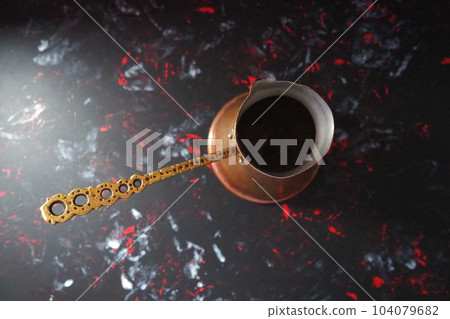 Balkan cezve for turkish coffee. A copper cezve on black surface. Antique bronze utensils. Carved metal handle with pattern. Black background with white, red and blue spots Balkan cezve for turkish coffee. A copper cezve on black surface. Antique bronze utensils. Carved metal handle with pattern. Black background with white, red and blue spots 104079682