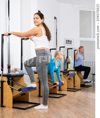 Relaxed young female standing and exercising with Pilates Wanda chair during group training in rehabilitation center Relaxed young female standing and exercising with Pilates Wanda chair during group training in rehabilitation center 104079825