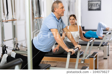Focused old man performing stretching Pilates exercises with reformer bed machine during group workout in gym Focused old man performing stretching Pilates exercises with reformer bed machine during group workout in gym 104079827