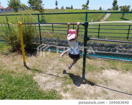 Back view of a child playing while hanging from a horizontal bar in a summer park 104083640