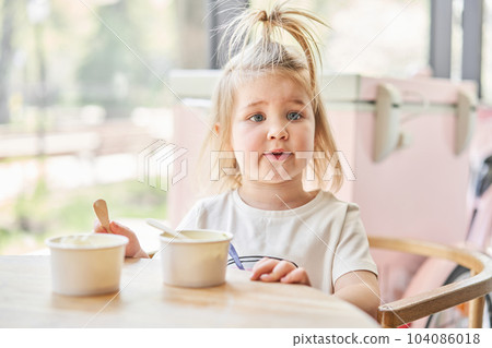 Cute little girl is eating ice cream. A beautiful charming baby girl sitting at a table in a cafe near the window. Children's cafe. The interior of an ice cream cafe. 104086018