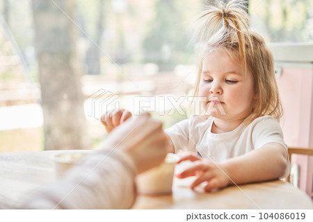 Cute little girl is eating ice cream. A beautiful charming baby girl sitting at a table in a cafe near the window. Children's cafe. The interior of an ice cream cafe. 104086019