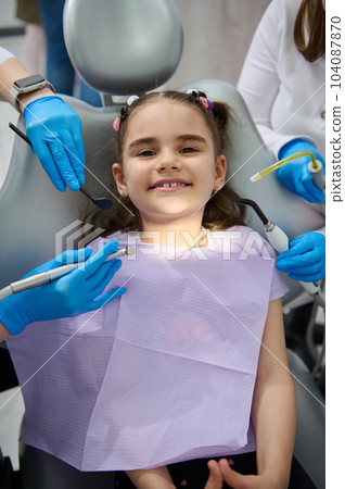 Top view. Hands of a dentist and assistant hold sterile dental instruments - dental mirror, probe, drill and saliva ejector above a smiling little patient at appointment in pediatric dentistry clinic Top view. Hands of a dentist and assistant hold sterile dental instruments - dental mirror, probe, drill and saliva ejector above a smiling little patient at appointment in pediatric dentistry clinic 104087870