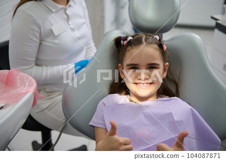 Smiling little girl at dentist appointment, sitting on dental chair, showing thumbs up after preventive dental check up in modern children's dental clinic. Pediatric dentistry. Prevention of caries 104087871