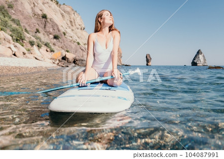 Woman sea sup. Close up portrait of happy young caucasian woman with long hair looking at camera and smiling. Cute woman portrait in a white bikini posing on sup board in the sea 104087991