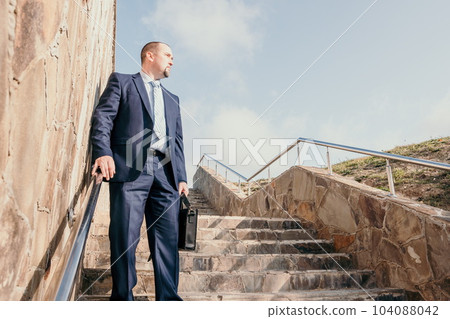 Confident middle age businessman with briefcase walking upstairs. Close-up of businessman wearing blue suit holding bag walking down the stairs Confident middle age businessman with briefcase walking upstairs. Close-up of businessman wearing blue suit holding bag walking down the stairs 104088042