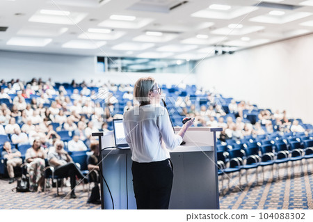Female speaker giving a talk on corporate business conference. Unrecognizable people in audience at conference hall. Business and Entrepreneurship event 104088302