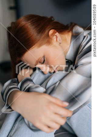 Close-up vertical portrait of sad millennial redhead female sitting on bed embrace knees lost in bad pessimistic thoughts suffer alone with closed eyes. Stressed young woman feel lonely desperate Close-up vertical portrait of sad millennial redhead female sitting on bed embrace knees lost in bad pessimistic thoughts suffer alone with closed eyes. Stressed young woman feel lonely desperate 104089589