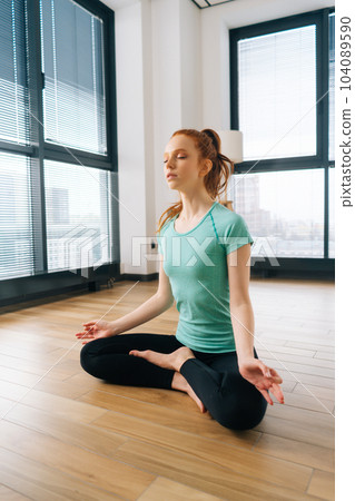 Vertical portrait of serene young woman meditating in lotus position sitting on floor with closed eyes in light living room enjoying meditation on background of window. Concept of healthy lifestyle. Vertical portrait of serene young woman meditating in lotus position sitting on floor with closed eyes in light living room enjoying meditation on background of window. Concept of healthy lifestyle. 104089590