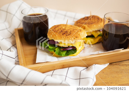 Big and tasty burgers lie on a wooden tray with a glass of cola. Wooden table and white checkered napkin. Close-up. 104091621