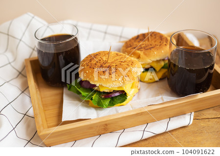 Big and tasty burgers lie on a wooden tray with a glass of cola. Wooden table and white checkered napkin. Close-up. Big and tasty burgers lie on a wooden tray with a glass of cola. Wooden table and white checkered napkin. Close-up. 104091622
