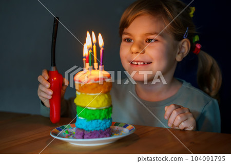 Happy little preschool girl celebrating birthday. Closeup of child with homemade rainbow cake, indoor. Happy healthy toddler blowing six candles on cake. Selective focus on cake 104091795