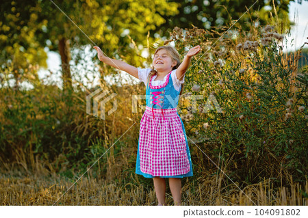Cute little kid girl in traditional Bavarian costume in wheat field. Happy child with hay bale during Oktoberfest in Munich. Preschool girl play at hay bales during summer harvest time in Germany. Cute little kid girl in traditional Bavarian costume in wheat field. Happy child with hay bale during Oktoberfest in Munich. Preschool girl play at hay bales during summer harvest time in Germany. 104091802