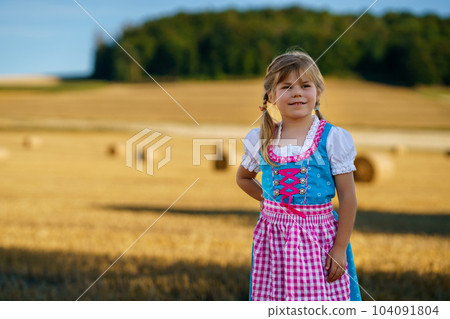 Cute little kid girl in traditional Bavarian costume in wheat field. Happy child with hay bale during Oktoberfest in Munich. Preschool girl play at hay bales during summer harvest time in Germany. Cute little kid girl in traditional Bavarian costume in wheat field. Happy child with hay bale during Oktoberfest in Munich. Preschool girl play at hay bales during summer harvest time in Germany. 104091804