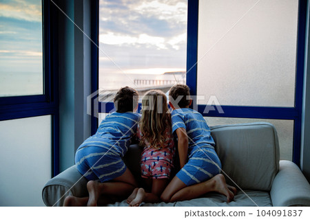 Three children watching sunrise on terrace or balcony. Two kids boys and preschool girl watch sun on horizon. Happy children relaxing near beach, family enjoying beautiful view. 104091837