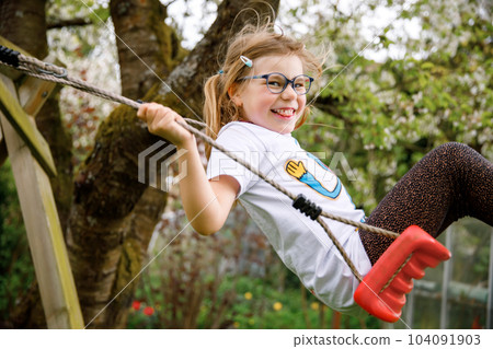 Happy little preschool girl with glasses having fun on swing in domestic garden. Cute healthy child swinging under blooming trees on sunny spring day. Kid laughing and crying. Happy little preschool girl with glasses having fun on swing in domestic garden. Cute healthy child swinging under blooming trees on sunny spring day. Kid laughing and crying. 104091903