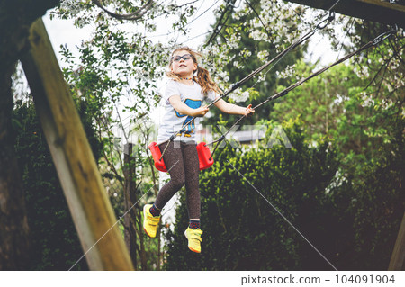 Happy little preschool girl with glasses having fun on swing in domestic garden. Cute healthy child swinging under blooming trees on sunny spring day. Kid laughing and crying. 104091904