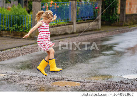 Happy little preschool girl wearing yellow rain boots and walking during puddles. Cute child in colorful clothes jumping into puddle, splashing with water, outdoor activity 104091965