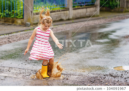 Happy little preschool girl wearing yellow rain boots and walking during puddles. Cute child in colorful clothes jumping into puddle, splashing with water, outdoor activity Happy little preschool girl wearing yellow rain boots and walking during puddles. Cute child in colorful clothes jumping into puddle, splashing with water, outdoor activity 104091967
