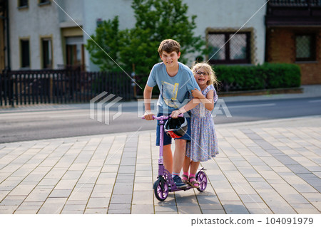 Preschool girl and school boy riding scooter in the city. Little sister and kid brother ride a roller. Kids play outdoors with scooter. Active leisure and outdoor sport for children 104091979