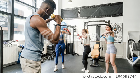 Young diverse ladies using dumbbells while training with instructor Young diverse ladies using dumbbells while training with instructor 104092744