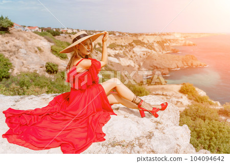 woman red silk dress sits by the ocean with mountains in the background, her dress swaying in the wind. 104094642
