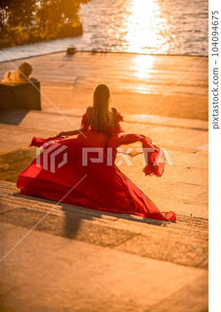 Sunrise red dress. A woman in a long red dress against the backdrop of sunrise, bright golden light of the sun's rays. The concept of femininity, harmony. 104094675