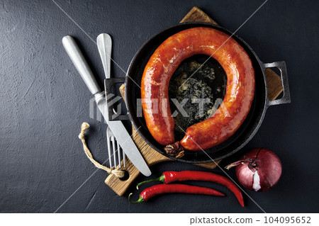 Fried sausage rolled into a ring on a cast iron pan with cutlery, a wooden cutting board, hot peppers and red onions on a black stone background Fried sausage rolled into a ring on a cast iron pan with cutlery, a wooden cutting board, hot peppers and red onions on a black stone background 104095652